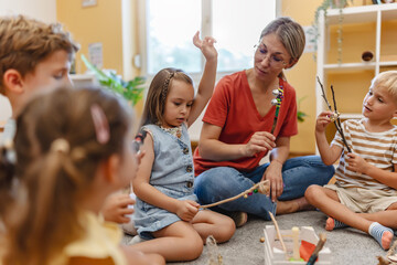 Preschool teacher leading a fun interactive activity with children sitting in a circle, raising hands and smiling in a bright classroom