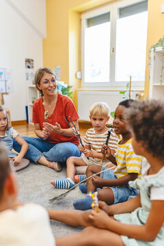 Preschool teacher leading a fun interactive activity with children sitting in a circle, raising hands and smiling in a bright classroom