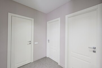 neutral-toned hallway features three white paneled doors with silver hardware. The walls are a light gray/lavender color, and the floor is tiled