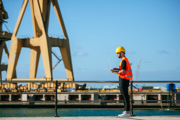 Naklejka premium Dock worker using laptop next to crane in a commercial harbor