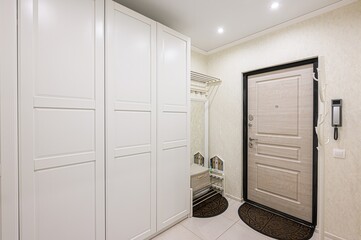 bright hallway features a white wardrobe, a door with a black frame, and a beige wallpaper.  There are rugs and a shoe rack near the door