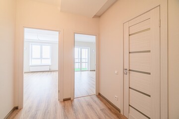 brightly lit hallway features wooden floors, neutral walls, and three doorways. One is a closed modern door, the others open to rooms with windows