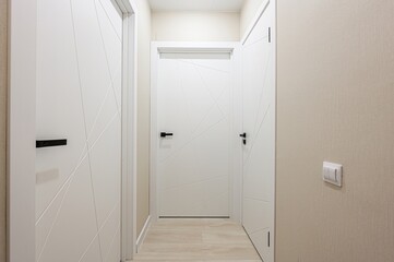 modern hallway featuring three white doors with geometric designs and black handles. The walls are light beige with a light switch