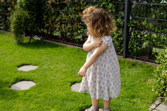 Cute toddler girl in a floral dress scratches her arm while playing outdoors in a sunny garden.