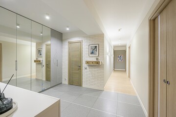 well-lit hallway with a mirrored wardrobe, wooden doors, and a white brick accent wall. Gray tile flooring transitions to a lighter wood floor in the distance