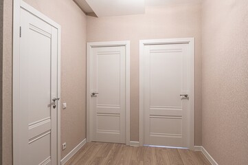 well-lit hallway featuring three white, paneled doors. The walls are covered in light beige wallpaper, and the floor is wood-toned laminate
