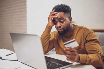 Worried African man using laptop and credit card while working remotely at his home. He is having financial problems.