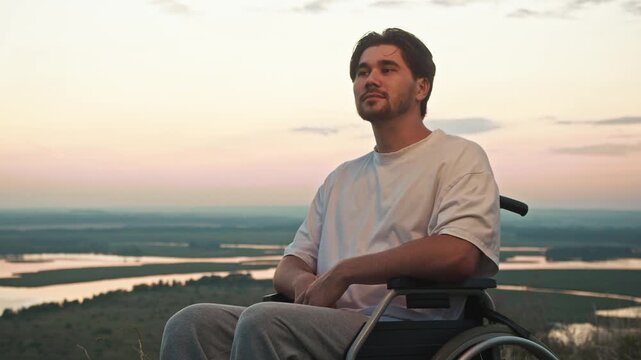 A man in a wheelchair admires the breathtaking sunset view from a hill with panoramic views of the green countryside and the distant riverbank.