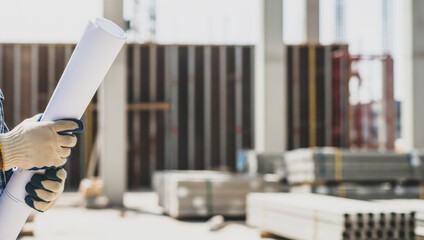 Person holding blueprints in construction site with building materials and unfinished walls,...