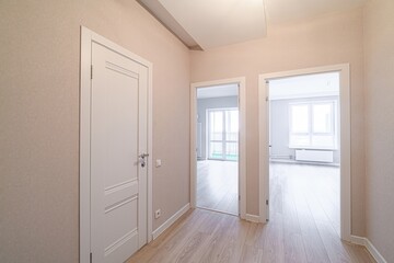 bright hallway with light beige walls and wood-look flooring. Three doorways are visible, two leading to sunlit rooms. A closed white door is on the left
