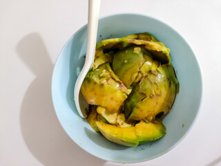 Top down view of a light blue bowl with fresh, ripe, sliced avocado and a spoon, a healthy food preparation.