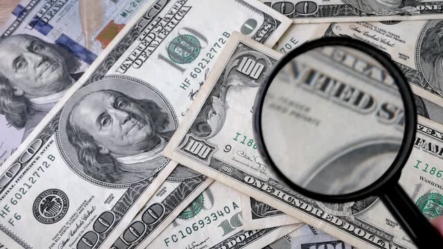 hand with magnifying glass checking 100 USA dollar banknote on money background, exchange money