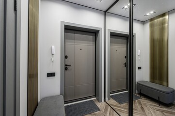 Modern hallway with gray walls, a door reflected in a large mirror, wooden flooring, and a gray bench. Wall decor includes gold vertical slats