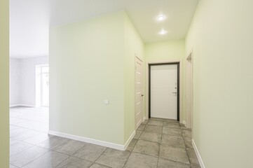 modern hallway with pale green walls, tiled floors, and white doors under recessed lighting. An open doorway leads to a bright room