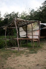 Rustic open shelter built from uneven wooden poles and a simple overhead lattice, partially covered with fabric. Structure stands on bare ground near tree line, suggesting a temporary © nicholasgeorgiou