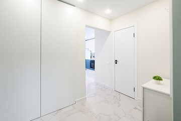 modern hallway with white walls, marble floor, and a glimpse of a kitchen. Minimalist design with a white door and cabinet