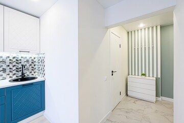 well-lit interior shows a modern kitchen area with blue cabinets, patterned backsplash, and an adjacent hallway featuring a white door and storage