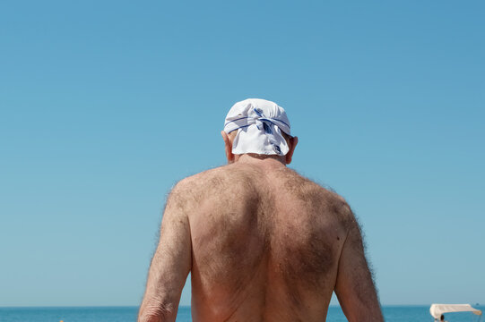 a man in a white headscarf stands with his back to the sea, sunbathing