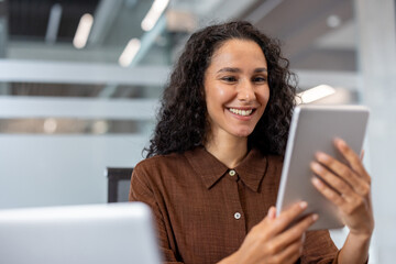 A cheerful woman interacts with a tablet in a bright, professional workspace. The contemporary setting and her smile convey positivity, connection, and productivity
