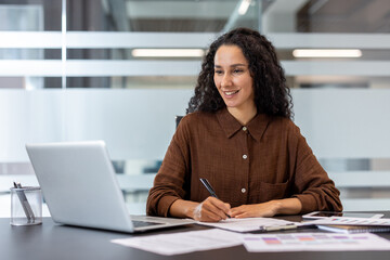 A professional woman sits at a desk, smiling while working on papers and using her laptop.