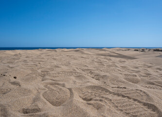 A beach with a person walking on the sand. The sky is blue and the sun is shining
