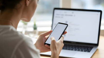 Focused woman multitasking with smartphone and laptop, engaging in digital workflow at home or office desk