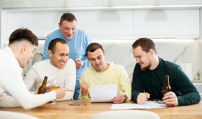 Fototapeta premium Happy friends drinking beer after solving their financial problems while sitting together at a home cooking table