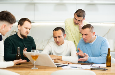 Group of men huddling around laptop at kitchen table, discussing problems and comforting distressed friend while gathering at home bachelor party