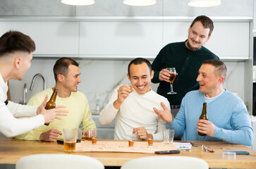 Cheerful young adults enjoying beer and wine immersed in board game with twist, where shot glasses of alcoholic drinks replacing traditional game pieces
