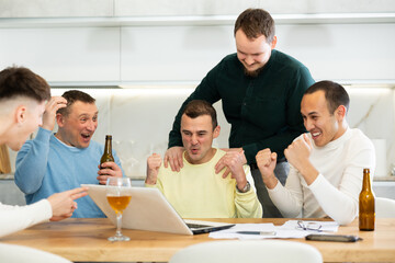 Fototapeta premium Excited friends gathered around laptop, celebrating successful investment made through online platform at casual home meeting with beer