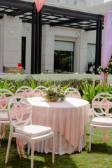 Elegant outdoor event setup with a round table covered in a peach tablecloth, surrounded by white chairs adorned with peach ribbons, set on green grass.