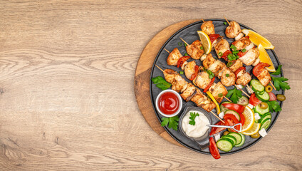 Grilled chicken skewers with fresh vegetable salad, lemon wedges, and dipping sauces on a round black platter. top view. copy space