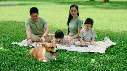 Smiling Asian family enjoying a casual picnic on the grass with their children and cheerful corgi dog, Selective focus on a pet