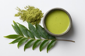 A small bowl of vibrant green matcha tea, beside a pile of matcha powder and a sprig of fresh green leaves, on a white background
