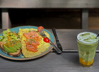 Two plates of avocado and red fish dishes served on a wooden table with a glass of matcha mango served with a metal straw. Healthy breakfast al fresco in natural light.

