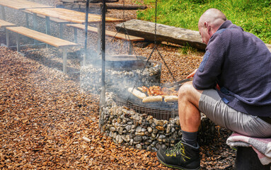 Man grilling sausages over an outdoor fire pit, surrounded by wooden benches and gravel, showcasing a rustic cooking experience in a natural setting