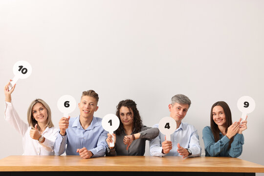 Panel of emotional judges voting with blank score signs at table against light grey background