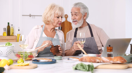 Elderly couple with laptop cooking together and drinking white wine at table indoors