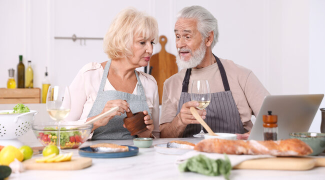 Elderly couple with laptop cooking together and drinking white wine at table indoors - Powered by Adobe