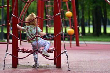 Red-haired toddler girl climbing a red rope ladder on a playground. Determined expression shows early childhood exploration, physical development, and outdoor play in a green forest park.