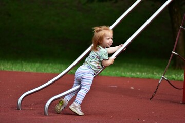 Red-haired toddler climbing up a shiny metal slide at a public playground. The child appears focused, gripping the bars with determination on a soft red rubber surface.