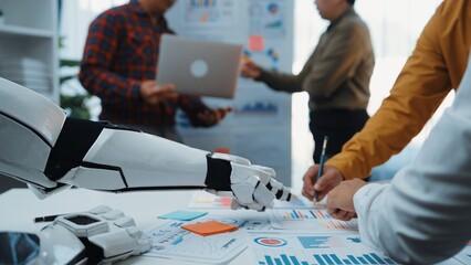A modern office scene showcasing a robot assistant engaging with professionals who discuss documents, laptops, and data analysis in a collaborative environment.Noogenesis