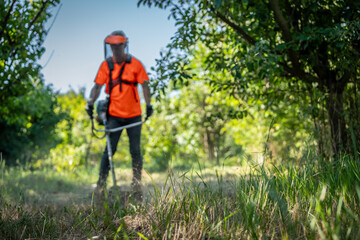 Close up of grass. In background is worker in special reflective clothing with a gasoline mower in his hand. A man with a trimmer mows grass. Man holding a brushcutter cut grass and brush.