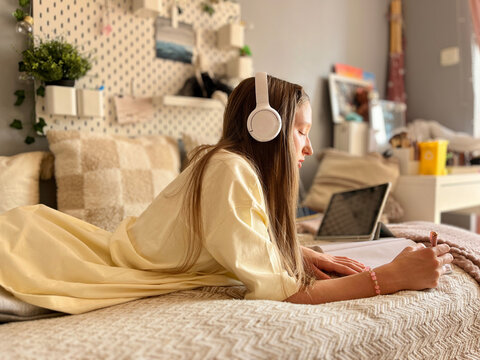 thoughtful teen girl lying on bed while writing in notebook and watching screen. Online learning and creative youth workflow in a home study environment. 