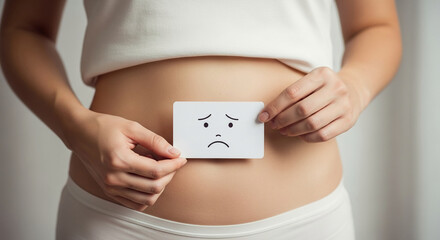 Woman holding sad face card on abdomen, representing discomfort or stomach issues, conveying feeling of illness or pain