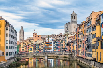 Colorful medieval houses along Onyar River in Girona, Spain, reflected in the water. Cathedral and basilica towers rise above, forming a picturesque Mediterranean cityscape