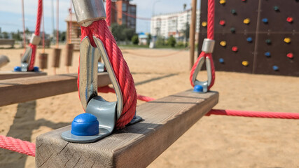 Close-up of wooden plank and red rope of suspended rope bridge on playground. Rope is secured with metal fasteners and blue plastic caps