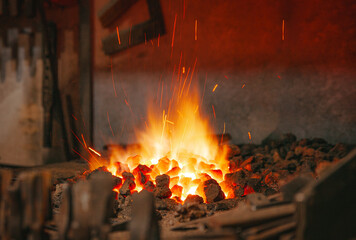 Bright flames and glowing embers illuminate a blacksmith's forge, with sparks flying upward against a warm, dark background. Tools and metal pieces are partially visible around the fiery center.