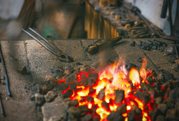Glowing coals radiate heat in a blacksmith's forge, surrounded by metal tools and ashes. The...