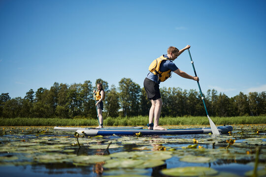 Caucasian young adult man and Caucasian young adult woman paddleboarding on lake covered with water lilies, both wearing life vests, standing on separate boards, trees in background - Powered by Adobe